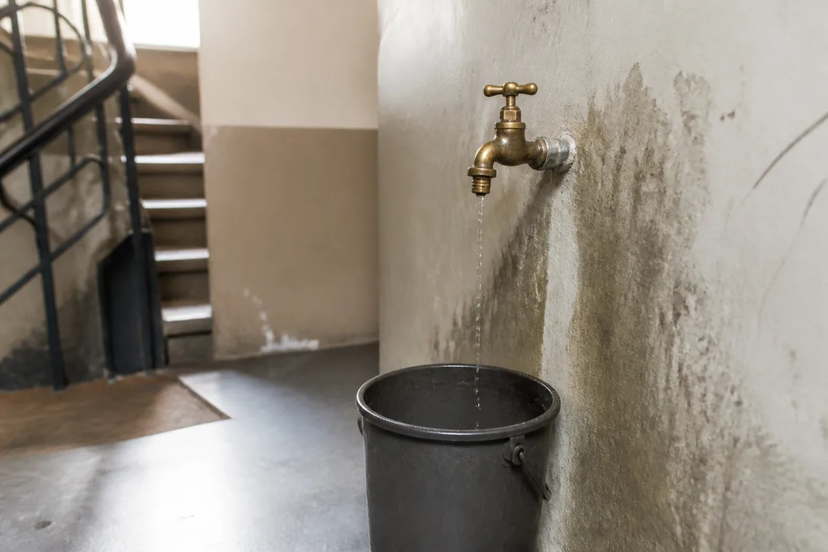 Robinet fuyant et trace d&rsquo;humidité sur un mur de cage d&rsquo;escalier, seau posé au sol